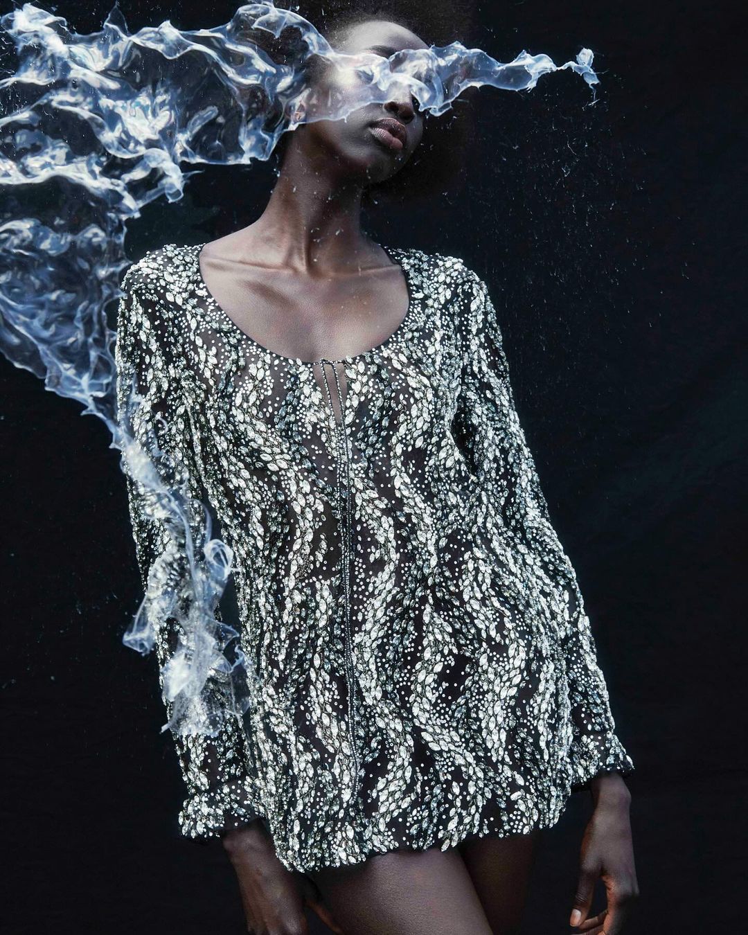 An African woman poses in a black and white patterned dress with a zipper in a dark studio setting with artistic water accents.