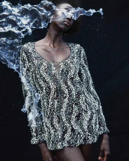 An African woman poses in a black and white patterned dress with a zipper in a dark studio setting with artistic water accents.
