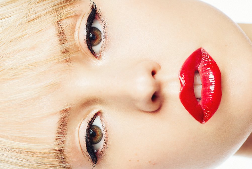 Close-up of a woman's face, featuring blonde hair, brown eyes with black eyeliner, and glossy red lips, set against a clean white background.