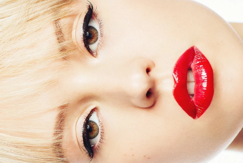 Close-up of a woman's face, featuring blonde hair, brown eyes with black eyeliner, and glossy red lips, set against a clean white background.