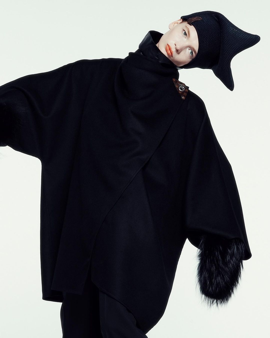 Fashion shot: woman in black cape and hat against white. High-fashion, editorial, indoor studio setting with theatrical costume elements.