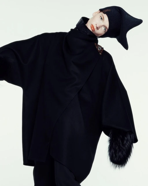 Fashion shot: woman in black cape and hat against white. High-fashion, editorial, indoor studio setting with theatrical costume elements.