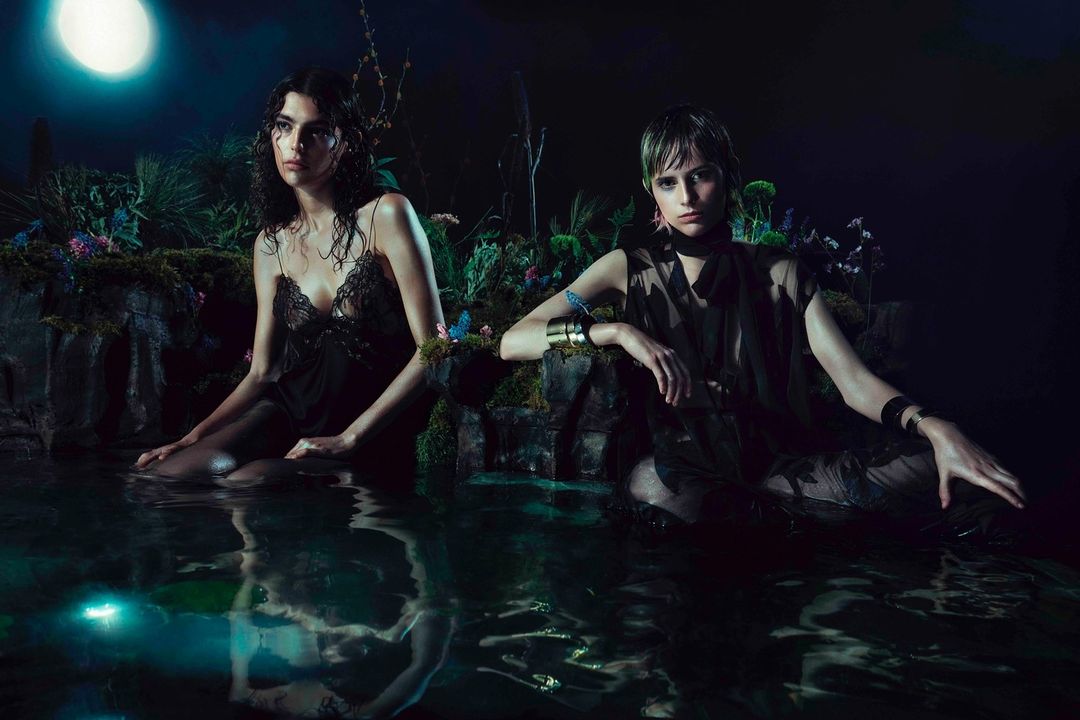 Two women in sheer dark clothing sit in shallow water at night, surrounded by lush plants and flowers under moonlight in a fashion editorial.