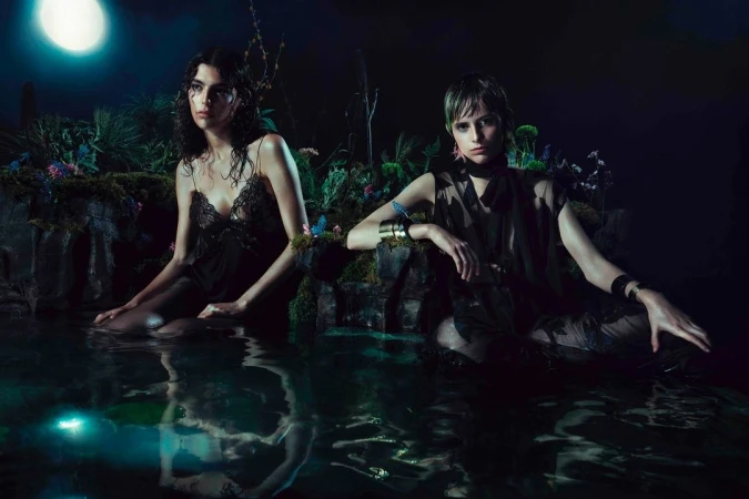 Two women in sheer dark clothing sit in shallow water at night, surrounded by lush plants and flowers under moonlight in a fashion editorial.