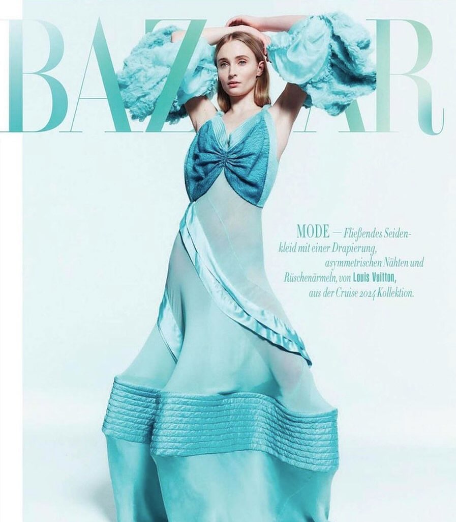 Editorial fashion shot: A fair-skinned woman in a ruffled aqua gown poses in front of the BAZAAR logo. The dress has dramatic sleeves.