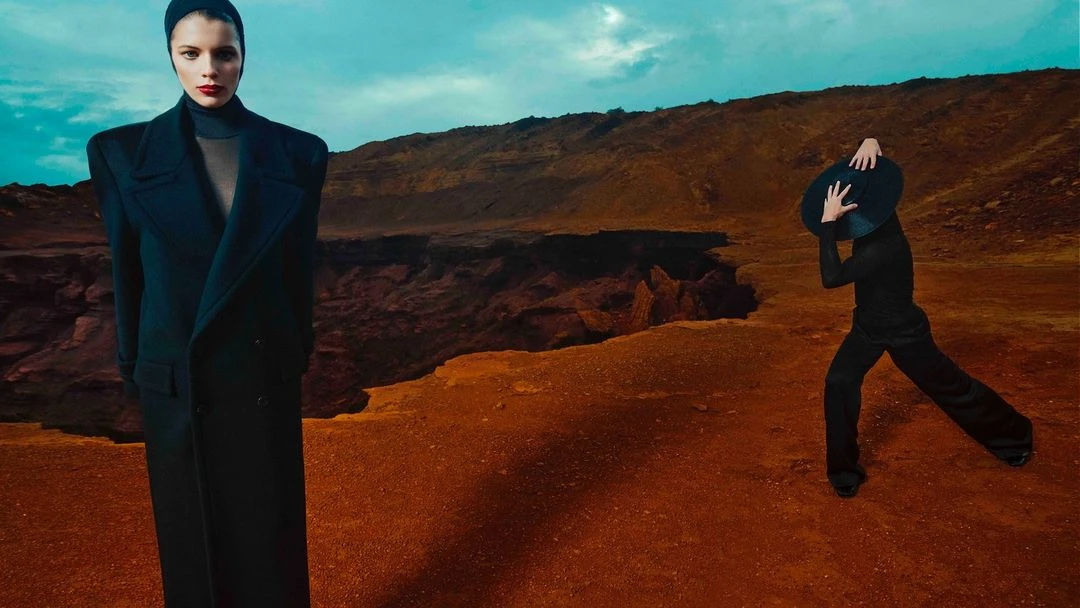 Two fashion models pose outdoors in black outfits against a surreal landscape with a blue sky. One holds a hat over their face.