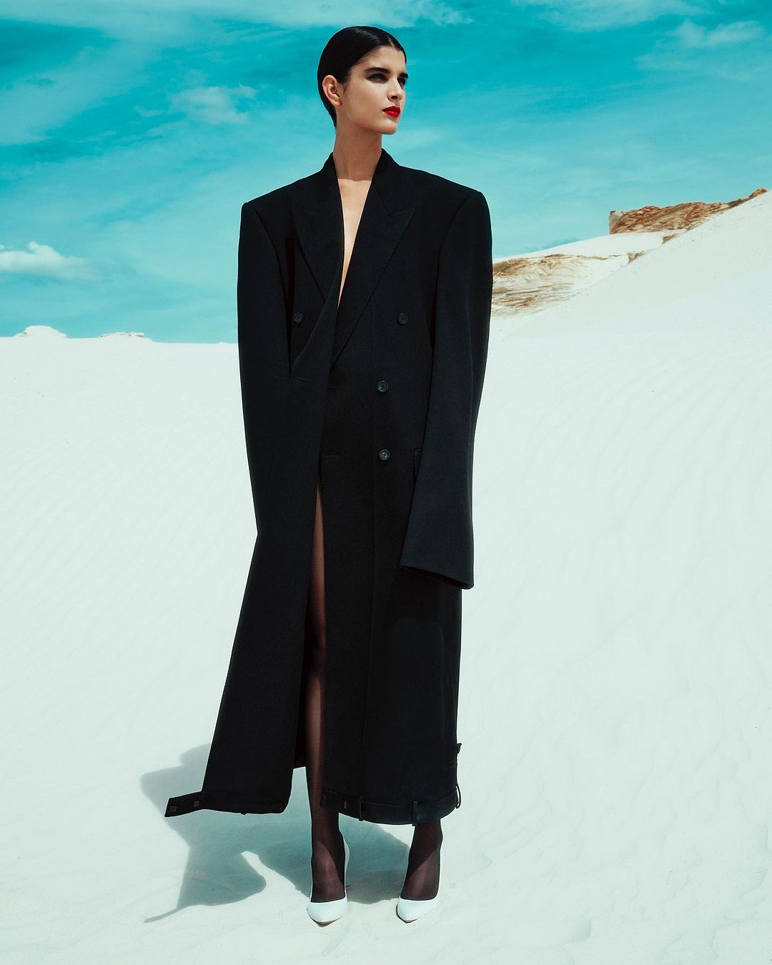 Elegant woman in a long black coat and white heels poses against a light blue sky and bright white sand dunes, creating a high fashion outdoor look.