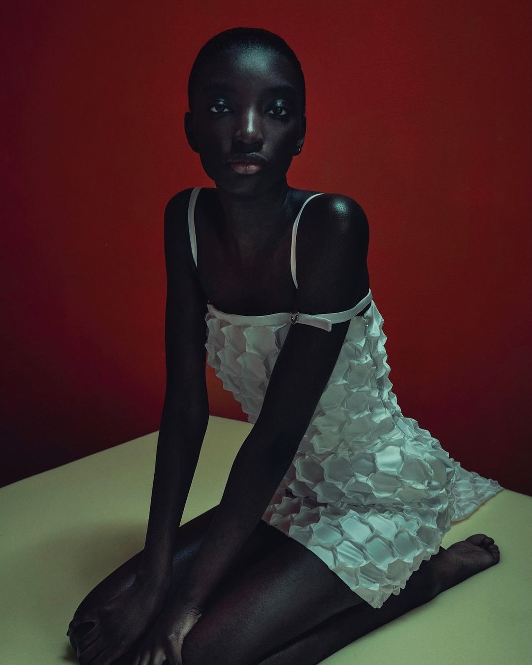 Elegant Black woman in a white dress poses kneeling in a photo studio against a red backdrop, showcasing fashion and beauty.