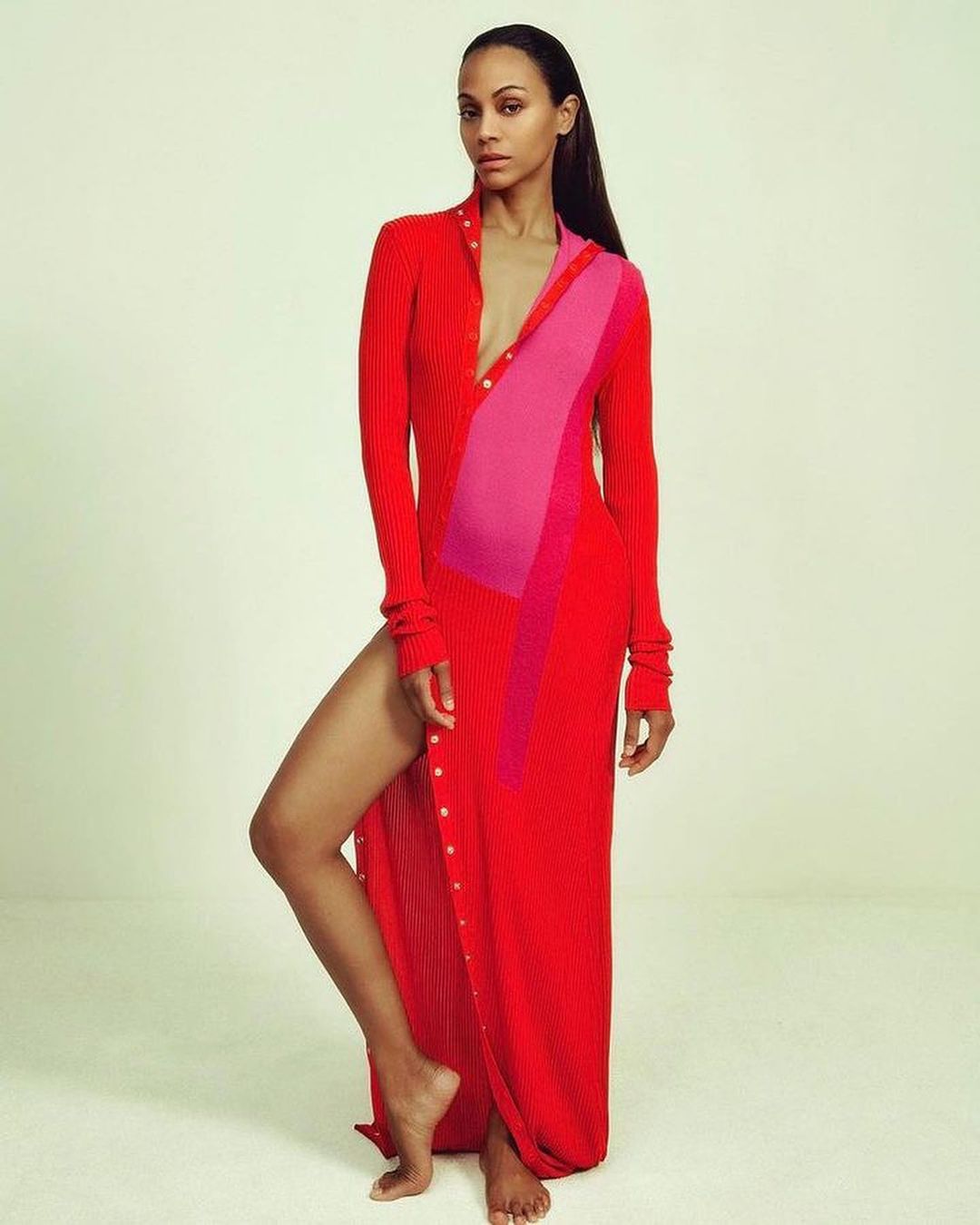 Zoe Saldana poses in a red button-down maxi dress with a pink geometric color block in a fashion studio shot on a white backdrop.