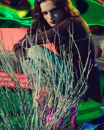 Model crouching outdoors behind sparse dry branches with vibrant green ground and shiny metallic background elements