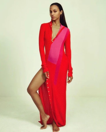 Zoe Saldana wearing a long red and pink ribbed dress with a high leg slit against a plain backdrop.