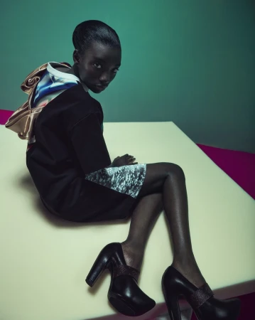 Black woman in a black dress and heels seated on a light surface against a green wall