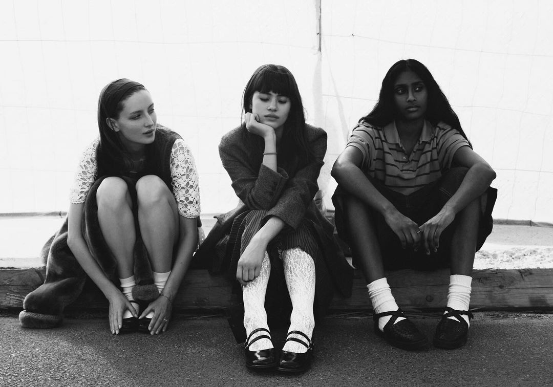 Three young women are sitting together on an unknown surface. The black and white photo emphasizes fashion details and a somber mood.