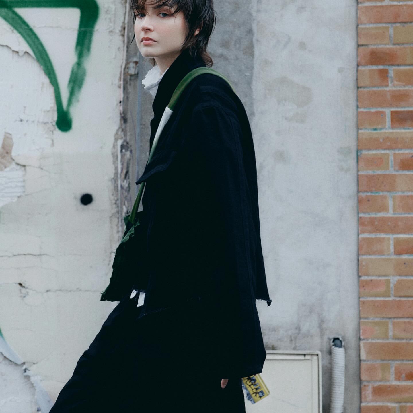 A young woman in a black coat stands against graffiti and brick walls, accented by a green bag, showcasing urban style in a neutral setting.