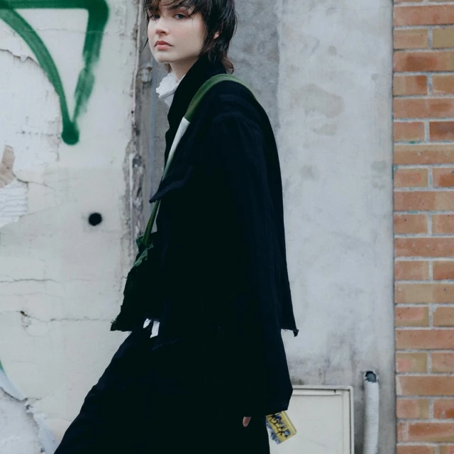 A young woman in a black coat stands against graffiti and brick walls, accented by a green bag, showcasing urban style in a neutral setting.