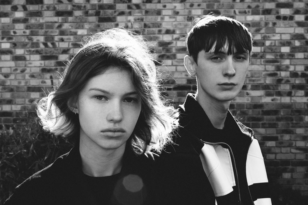 Black and white portrait of a young adult couple posing against a brick wall backdrop in outdoor setting with natural lighting.