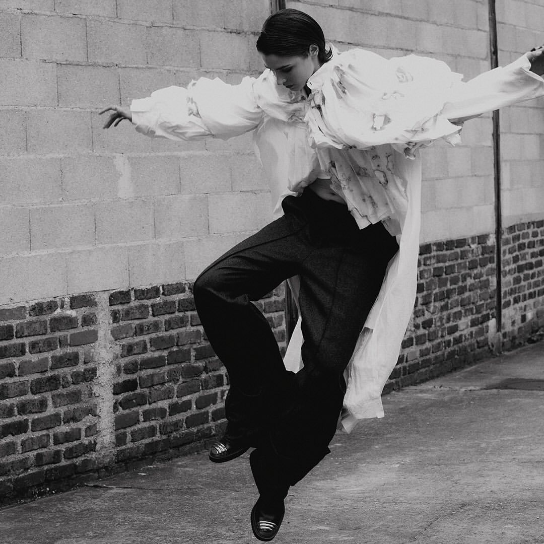 A woman in a long white shirt and dark pants is jumping in the air in front of a brick wall, captured in a black and white image.