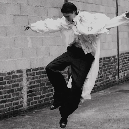 A woman in a long white shirt and dark pants is jumping in the air in front of a brick wall, captured in a black and white image.