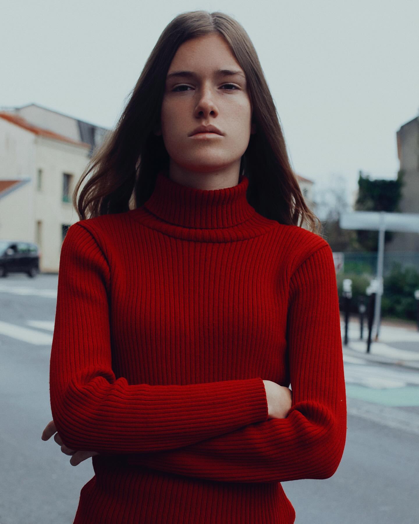 Portrait of a young adult woman with long brown hair wearing a red turtleneck sweater and arms crossed, posing outdoors on a city street.