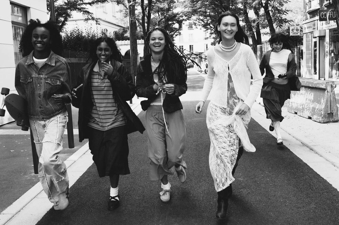 Five young women are walking on a street with buildings and trees in the background, creating a casual fashion scene in black and white.