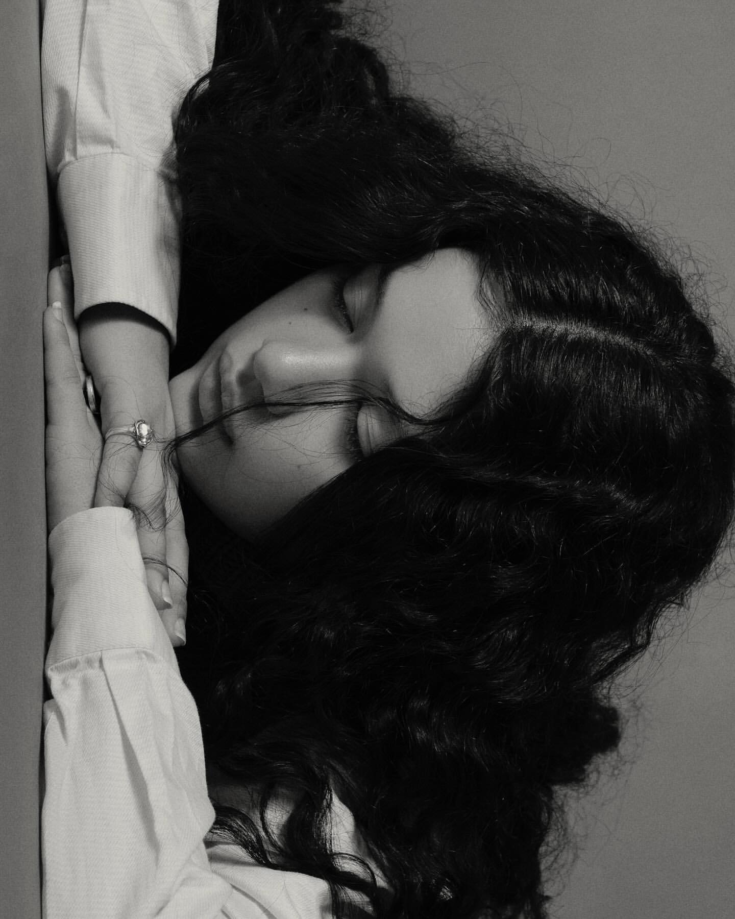 Monochrome portrait of a young woman with long, dark, wavy hair, wearing a collared shirt, gazing downward in a serene, artistic style.