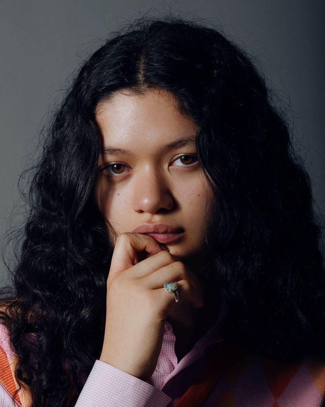 Close-up portrait of a young adult female with dark, curly hair wearing a patterned sweater, holding her hand to her mouth.