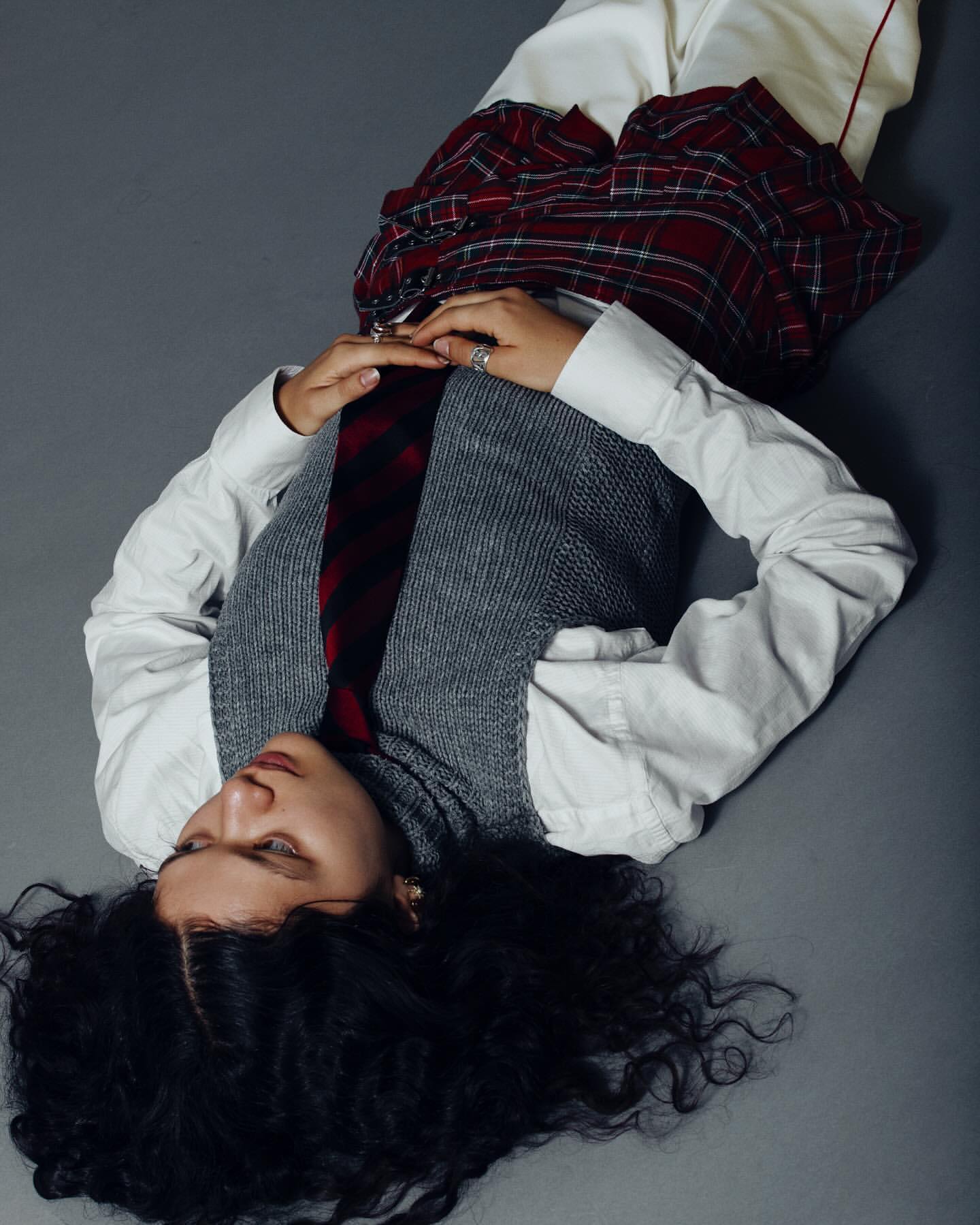Fashion shot of an adult woman lying on a gray backdrop wearing a white shirt, gray vest, plaid skirt, and dark tie.