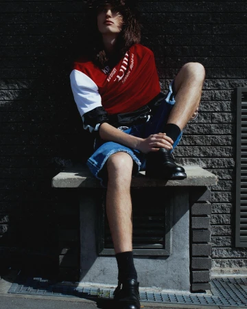 Model with long dark hair seated on a concrete structure wearing a red, white, and black jersey and denim shorts.