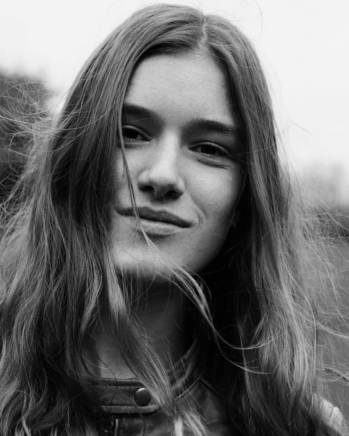 Portrait of a young adult woman with long hair, smiling slightly, captured in a close-up shot with a blurred outdoor background in black and white.