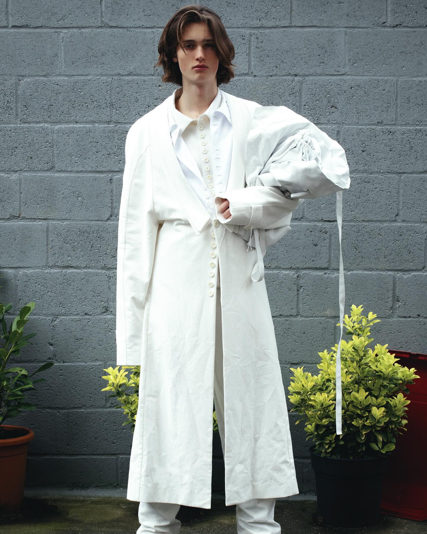 A young adult male posing in a long white coat, white pants, and a white shirt in an outdoor setting against a grey brick wall.