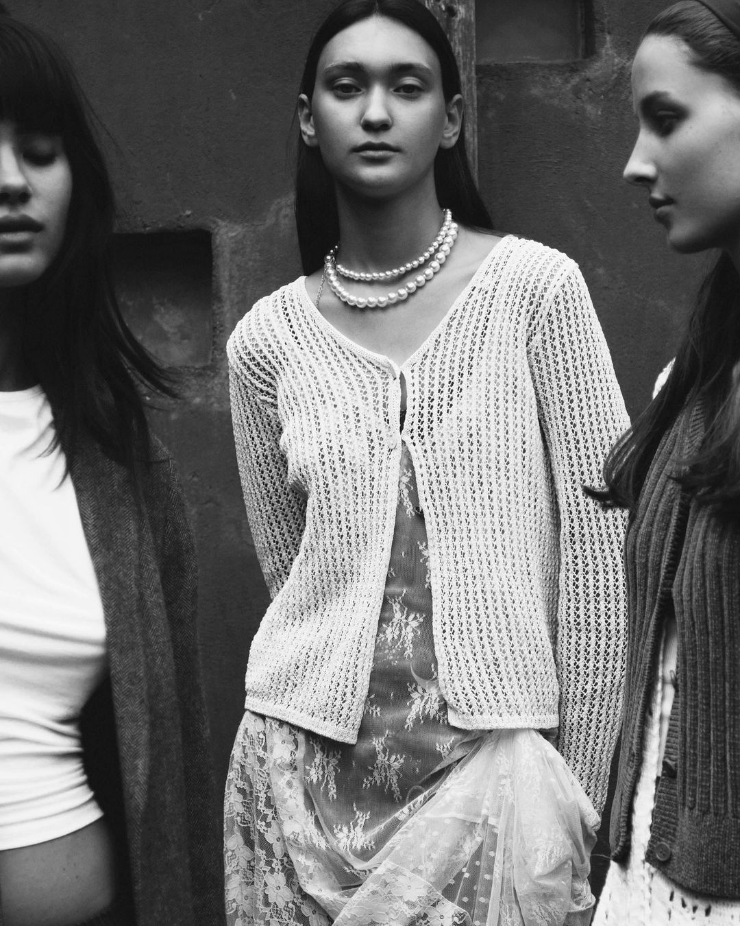 Three women in vintage fashion stand against a gray wall, wearing knit cardigans, lace dresses, and pearl necklaces in a monochromatic shot.