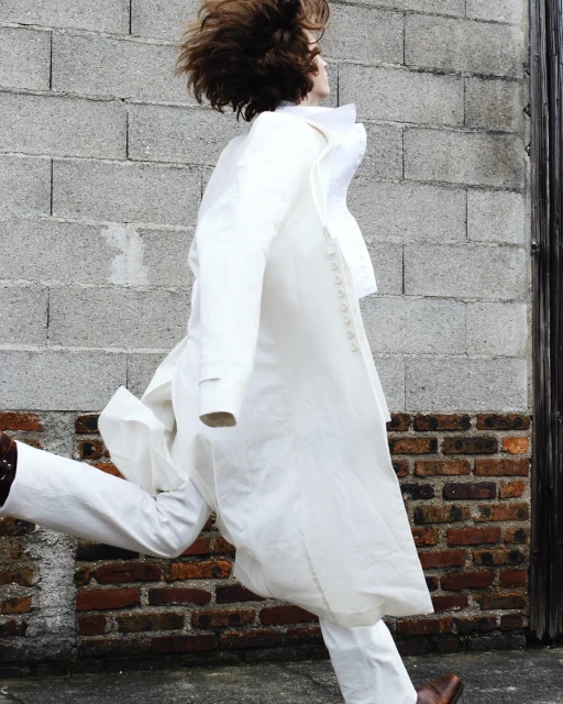 Fashion shot of a person with wind-blown hair, wearing a long white jacket and pants, jumping in front of a brick and concrete wall.