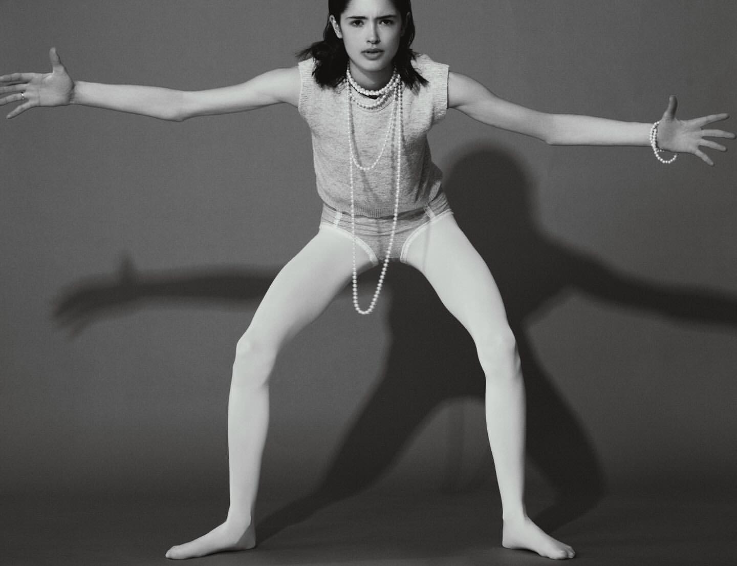 Young woman in a grey shirt, white tights, and pearls poses with outstretched arms in a studio against a grey backdrop with shadows.