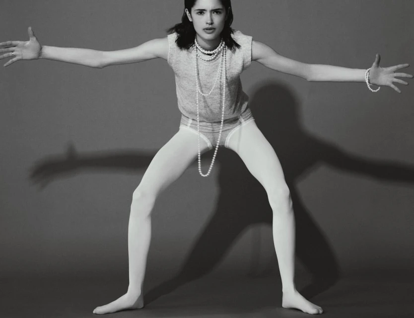 Young woman in a grey shirt, white tights, and pearls poses with outstretched arms in a studio against a grey backdrop with shadows.