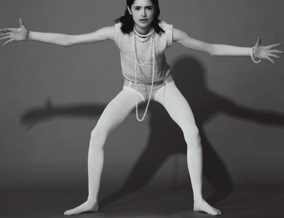 Young woman in a grey shirt, white tights, and pearls poses with outstretched arms in a studio against a grey backdrop with shadows.