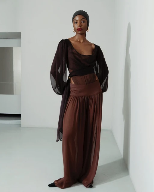 Fashion editorial shot of an African-American woman in brown sheer blouse, wide-leg chiffon pants, and a head covering, against a white wall.