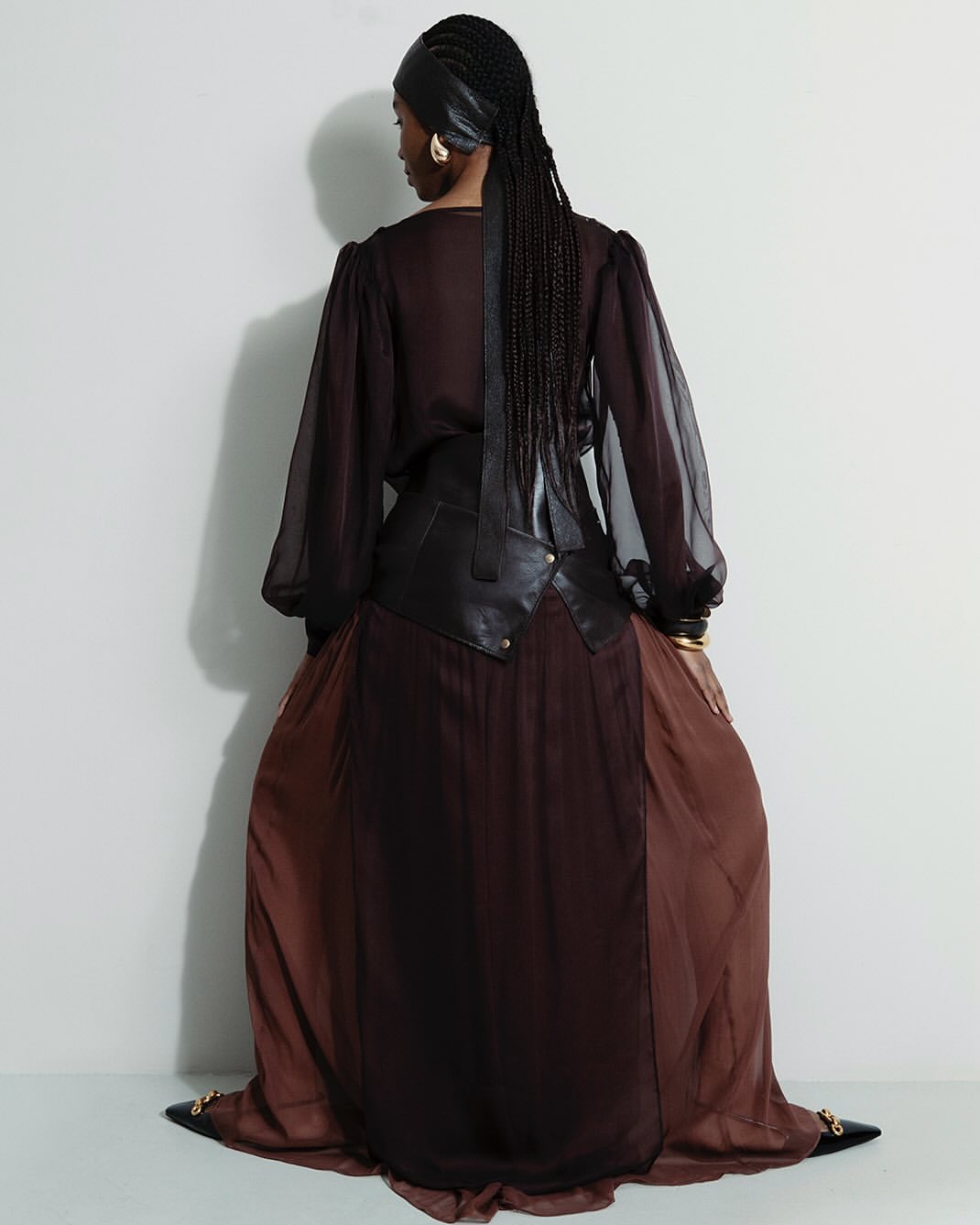 A woman with dark braided hair is facing away from the camera, wearing a brown dress and black leather accents against a white backdrop.