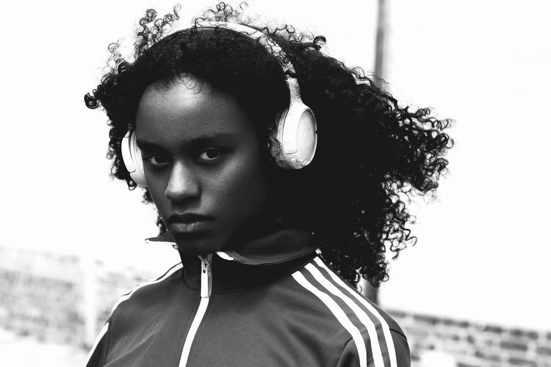 Monochrome portrait of a serious young Black woman with curly hair and headphones, wearing a striped track jacket against a bright background.