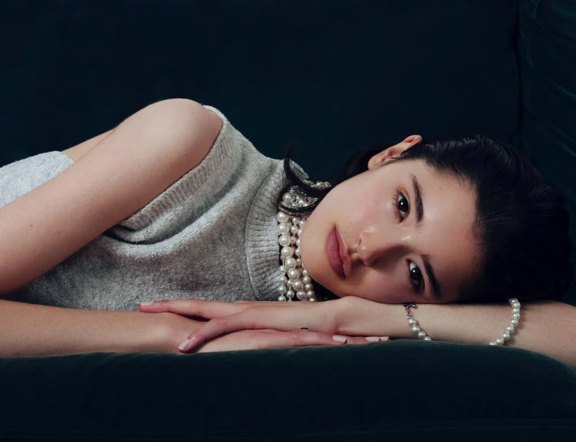 Portrait of a woman lying on a dark green velvet surface, adorned with pearl jewelry, wearing a light grey sweater in a studio setting.