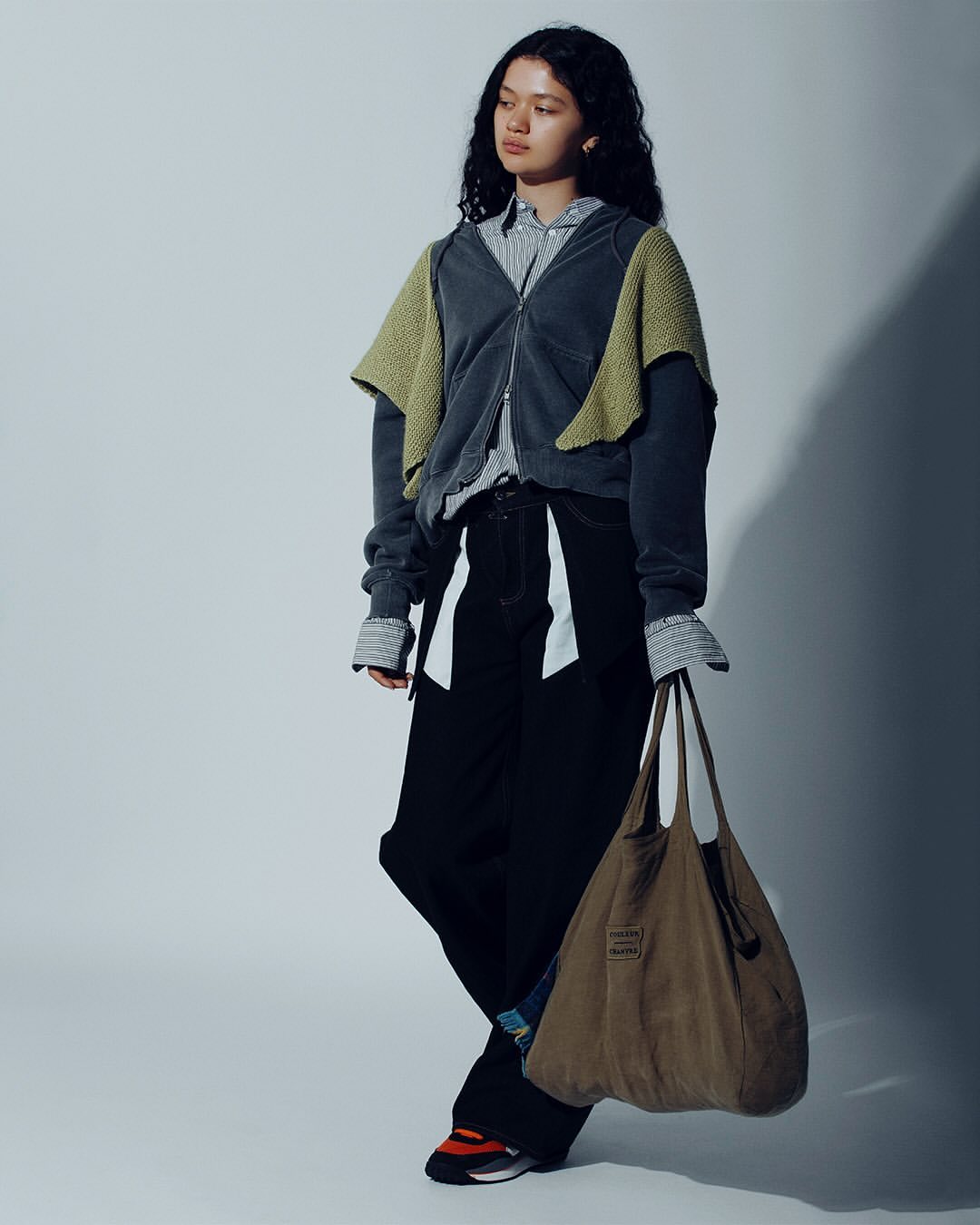 A young adult woman poses in a gray zip-up hoodie, black pants, and sneakers, holding a brown tote bag in a studio shot.