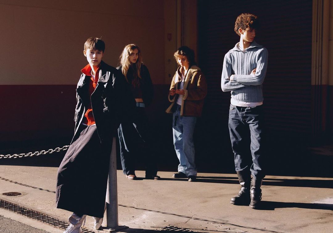 Four young adults stand in sunlight against a metal building, dressed in contemporary clothing. They are in an urban setting near an industrial area.