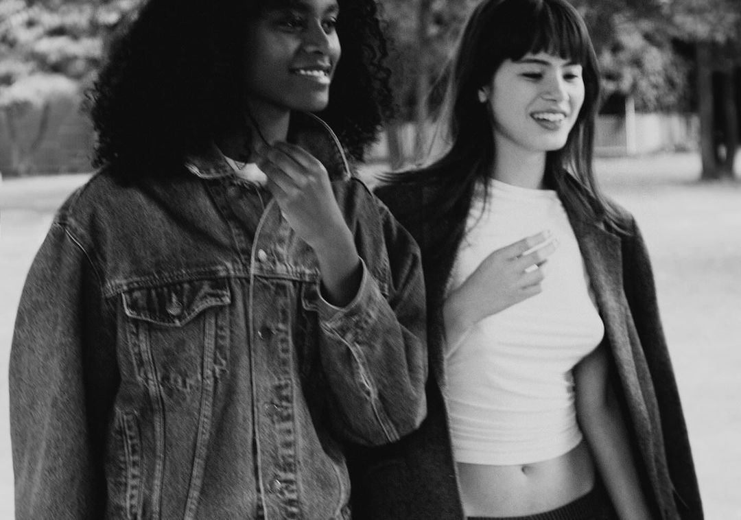 Two young adult women pose in a black and white outdoor portrait. One wears a denim jacket, and the other is in a sweater and coat.
