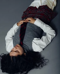 Model lying on grey studio floor wearing grey sweater vest, white shirt, plaid skirt, and striped tie by Soliou Ligali