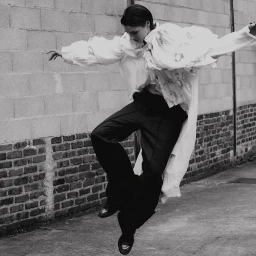 Model in motion wearing a white ruffled blouse and dark trousers jumping against a brick wall in Paris