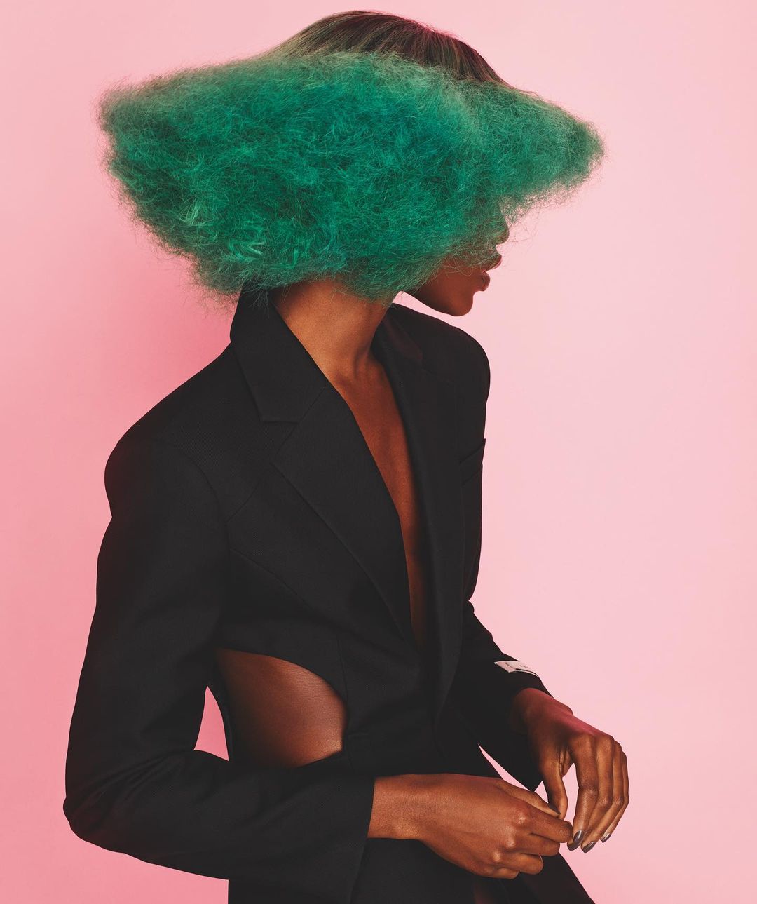 Studio fashion portrait of a Black female adult model wearing a black blazer with green cloud-like hair covering her face against a pink background.