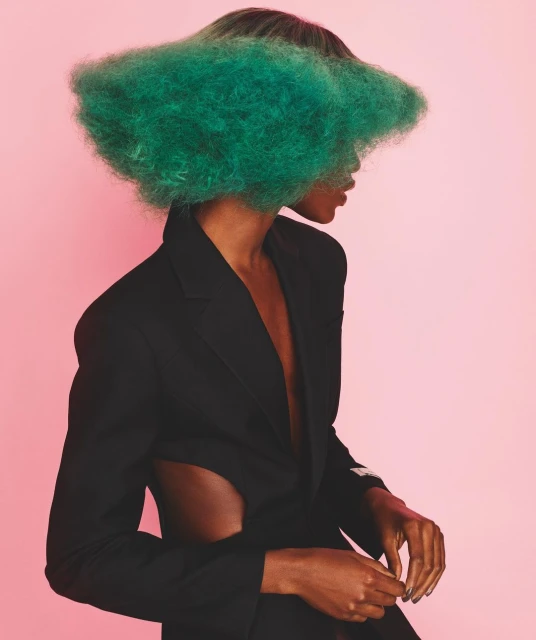 Studio fashion portrait of a Black female adult model wearing a black blazer with green cloud-like hair covering her face against a pink background.