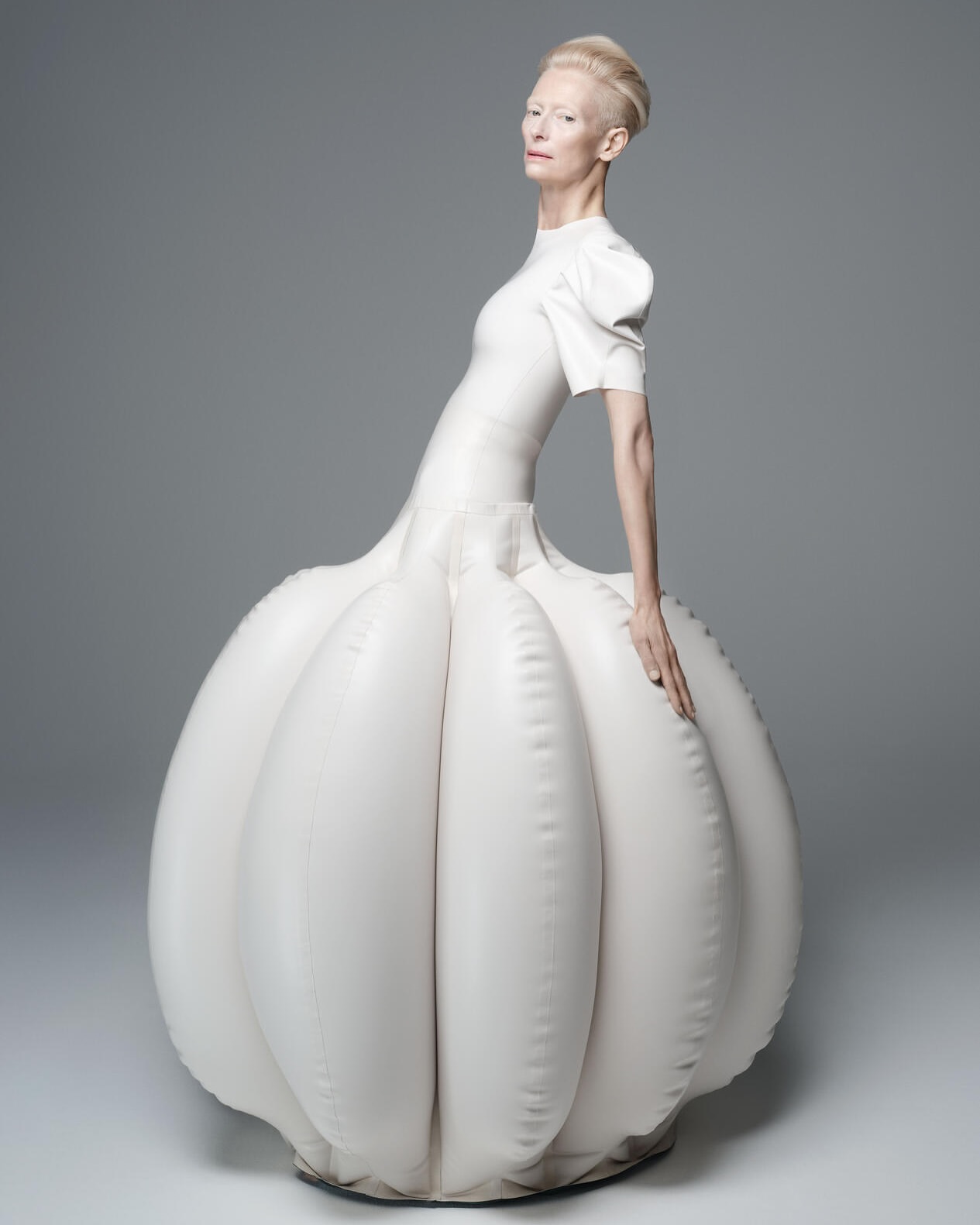 Tilda Swinton wearing an exaggerated voluminous white sculptural gown against a seamless grey studio background