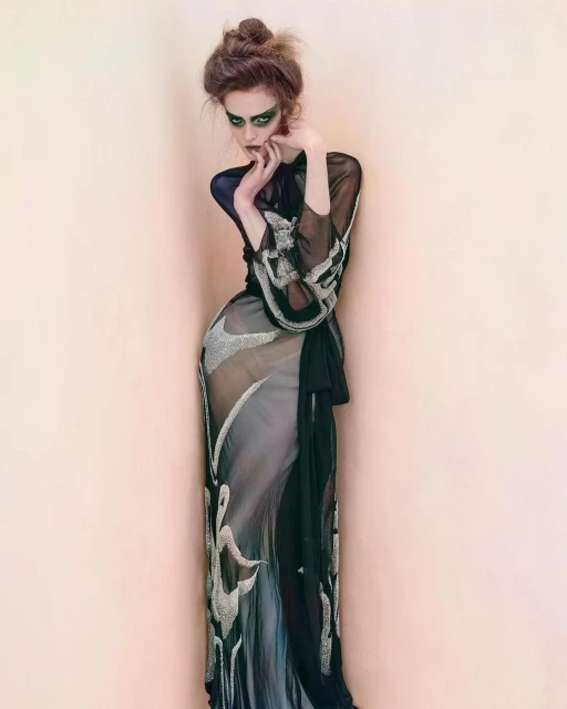 A woman in a sheer gown poses against a peach wall, featuring dramatic makeup, a high bun hairstyle, and hands near her face.