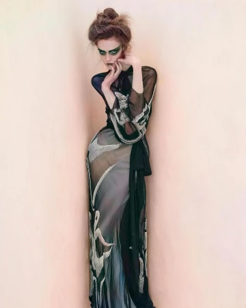 A woman in a sheer gown poses against a peach wall, featuring dramatic makeup, a high bun hairstyle, and hands near her face.
