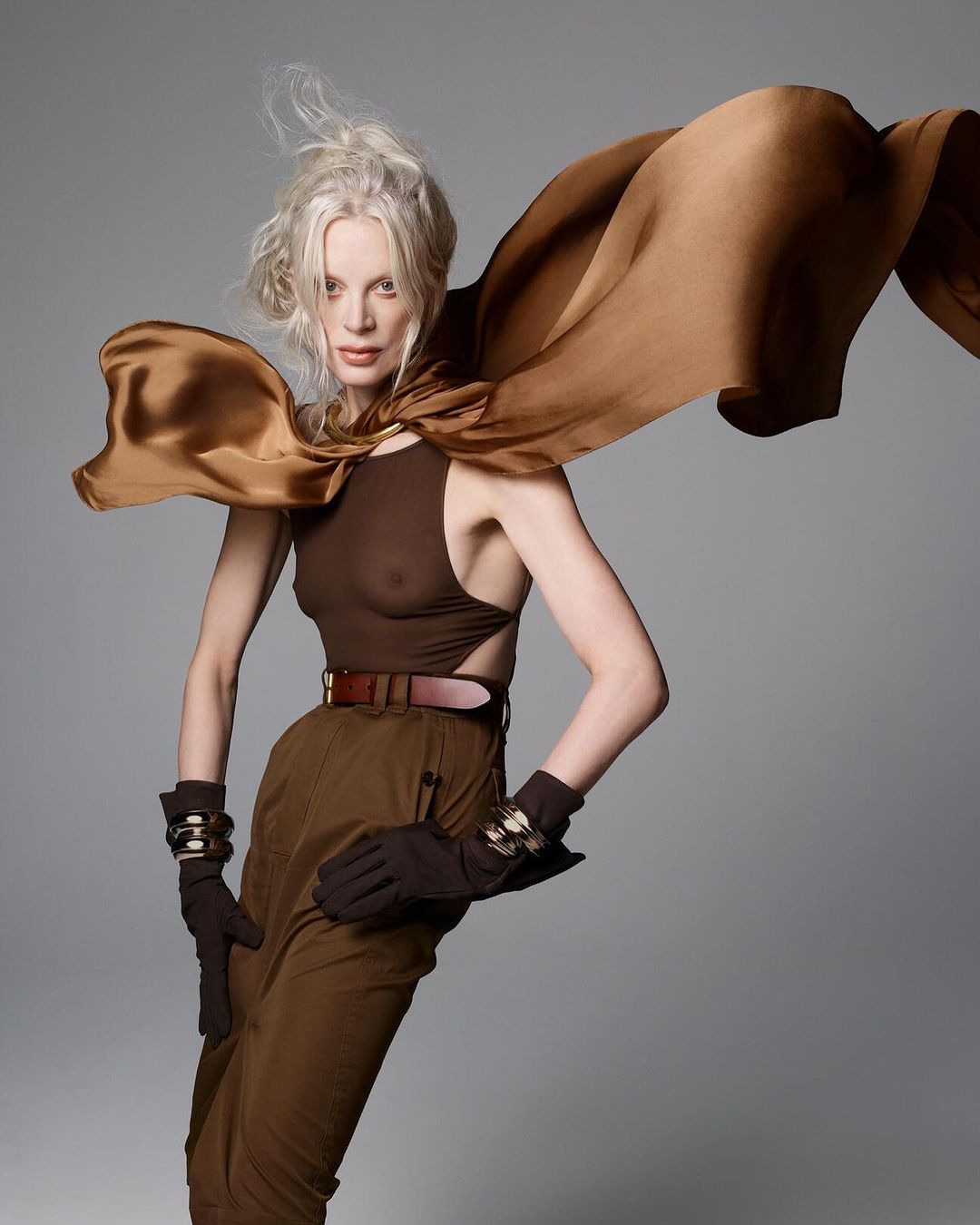 Fashion shot of a stylish, mature, blonde woman in a brown bodysuit, pants, and cape, striking a pose against a neutral gray backdrop.
